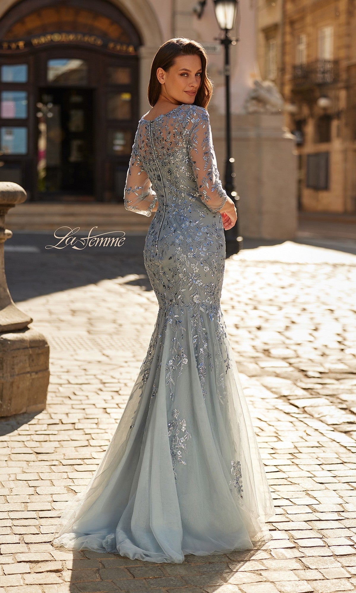 A woman stands outdoors on a cobblestone street wearing the La Femme 31799 long formal dress, a light blue mermaid gown with long sleeves, fitted bodice, flared skirt, and floral beaded embroidery.